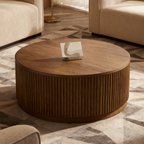 Round wooden coffee table in a living room setting with beige sofas and a patterned rug.